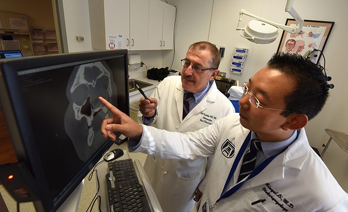 Two men with dark hair in white jackets, pointing at screen with skull xray