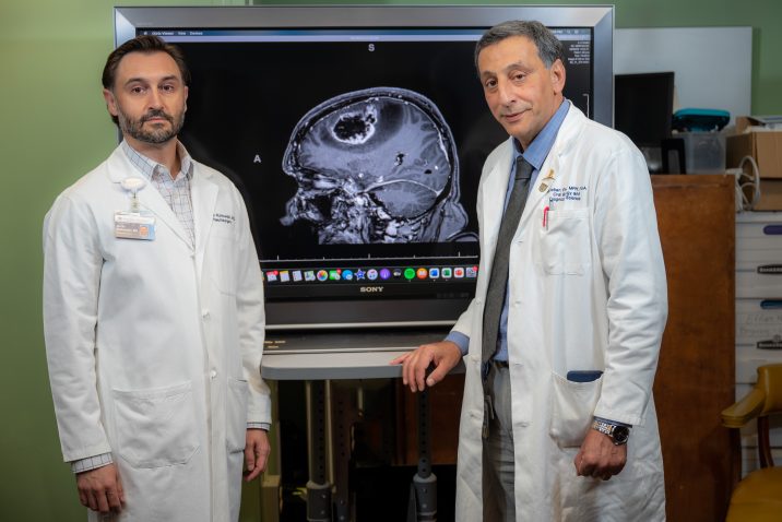 Two men in white coats stand in front of a scan showing a brain tumor