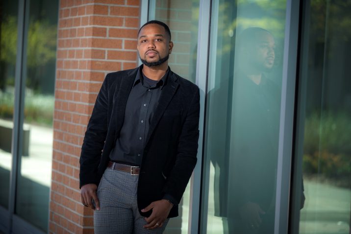 Man in black coat and shirt leans against building