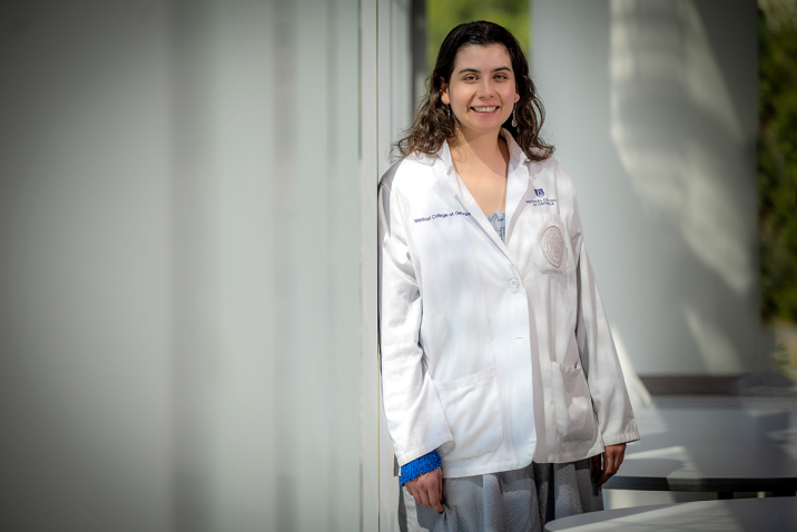 Woman wearing doctor's lab coat stands against a column while smiling for a picture.
