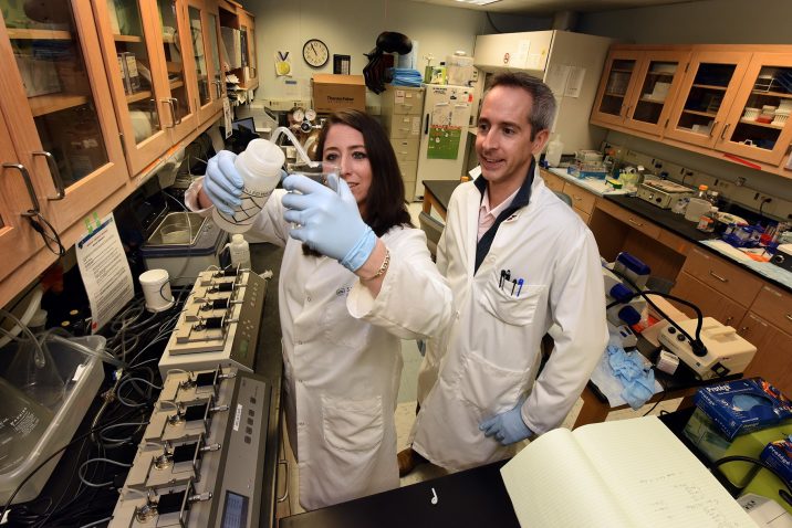 Woman with gloves pours liquid into beaker while man looks on