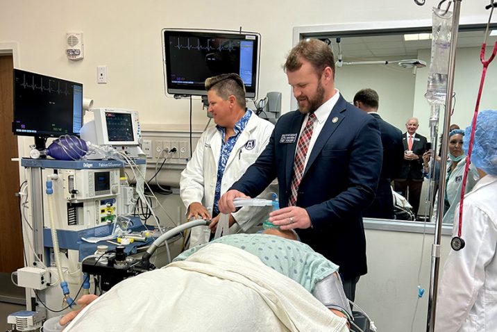 A man in a suit stands at the head of a gurney holding an anesthesia mask to a dummy patient while a woman in a lab coat watches a screen to his left