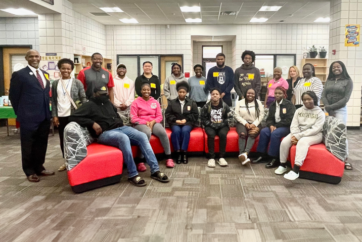 A group of students and teachers and administrators pose for a photo on and around a couch inside a school library