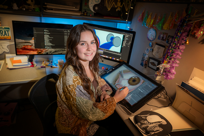 A female college student sits in a cubicle with several computer monitors around her. She is holding a stylus and there is an illustration of a human eye on one of the screens.