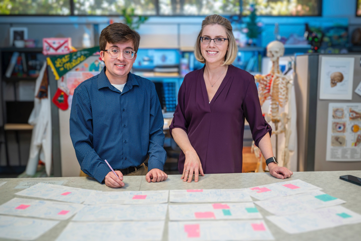 A college man and a professor look over mock-ups of a children's book spread over a table top.