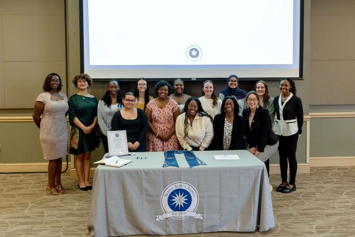 Female first-generation scholars stand gathered in front of an Alpha Alpha Alpha table.