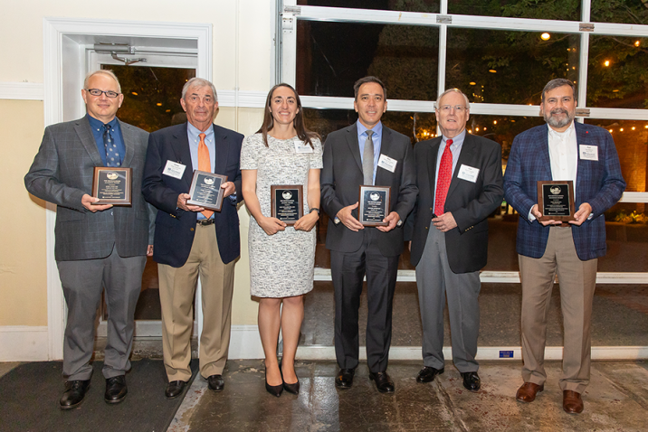 Six people, including five men and one woman, stand in front of a large group of people. Five of the people are holding up honorary plaques.