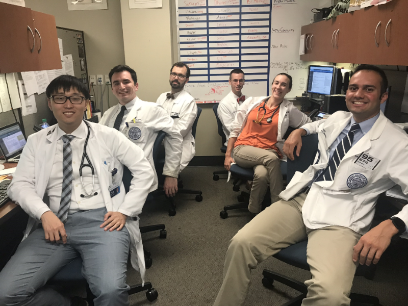 Six residents in white coats sitting in chairs in front of computers