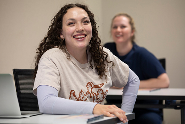 Female student with curly brown hair smiles from seat in classroom